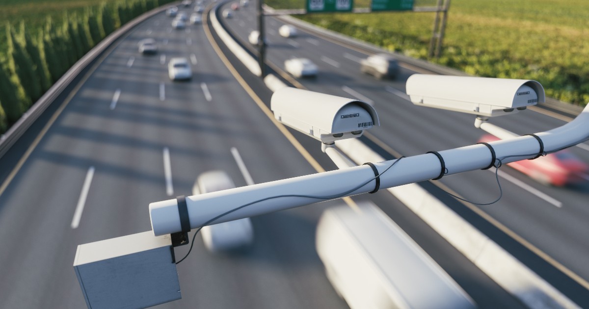 Imaginea prezintă camere de supraveghere și monitorizare a traficului montate pe o structură metalică deasupra unei autostrăzi aglomerate.