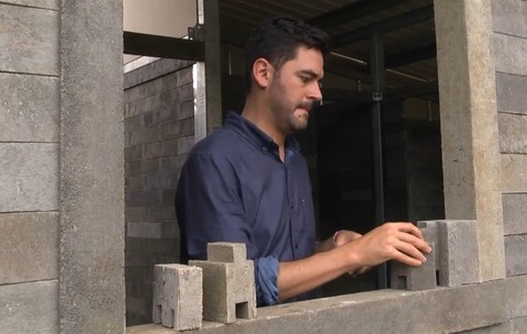 Imaginea prezintă un bărbat îmbrăcat în cămașă albastră care asamblează blocuri modulare din plastic reciclat pe glafurile unei ferestre în cadrul construcției unei case.