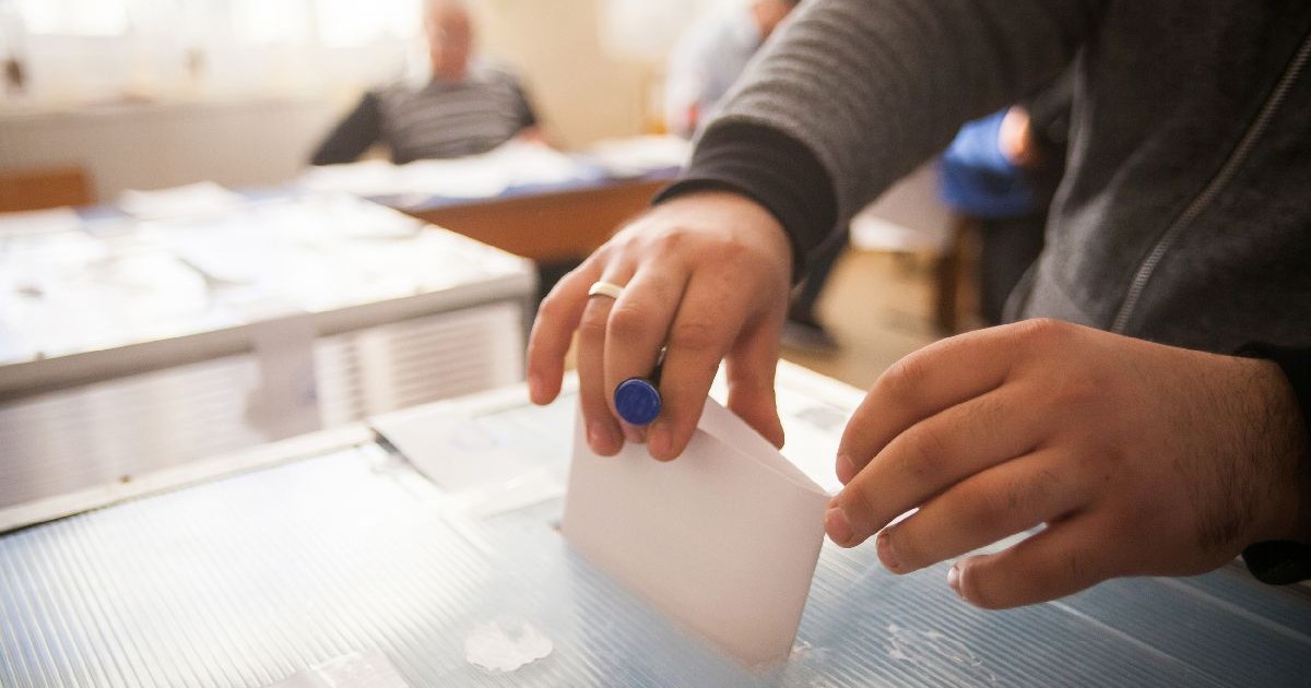 O persoană introduce buletinul de vot într-o urnă, la o secție de votare. În fundal se văd persoane estompate într-o sală.