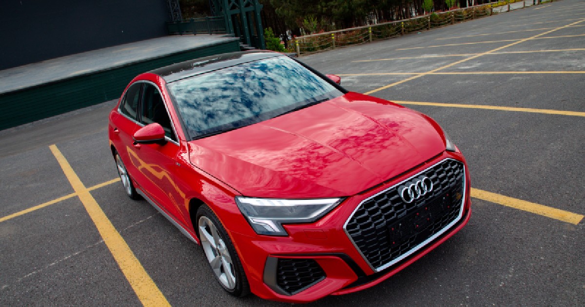 Imaginea prezintă un autoturism Audi roșu cu plafon negru parcat într-o zonă marcată cu linii galbene, având în fundal o structură de tip scenă și o zonă cu arbori.