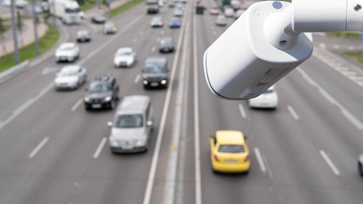 Autostradă aglomerată, cu mai multe benzi pe sens. În prim-plan, în partea dreaptă a imaginii, se vede o cameră de supraveghere rutieră montată deasupra carosabilului, orientată către traficul auto.