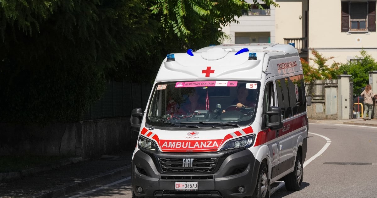 Imaginea prezintă o ambulanță văzută printr-un efect de suprapunere sau reflexie, cu luminile albastre aprinse, sugerând o intervenție de urgență. În fundal se observă siluete de persoane și elemente distorsionate, ceea ce creează o atmosferă tensionată și dramatică.