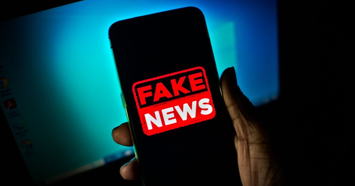 Un om ține în mână un telefon cu ecranul negru pe care apare mesajul roșu Fake News, iar în fundal se vede ecranul unui laptop, într-o cameră întunecată