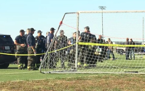 Polițiști adunați pe un teren de fotbal în spatele unei porți, într-o zonă izolată cu bandă galbenă de protecție a poliției, lângă un vehicul de intervenție.