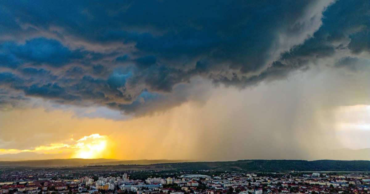 Vedere panoramică deasupra unui oraș, peste care se extind nori întunecați de furtună ce aduc o perdea densă de ploaie.