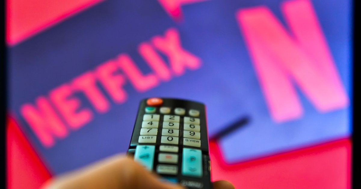Imagine creativă cu mâna unei persoane, care ține o telecomandă îndreptată către un televizor pe care este afișat logo-ul Netflix