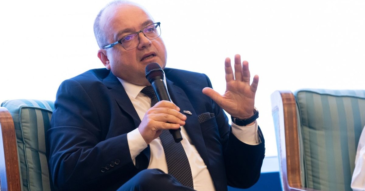 Profesorul Valentin Naumescu vorbește la microfon și explică ceva cu mâna, în timpul unei dezbateri. În spate, are un fond alb
