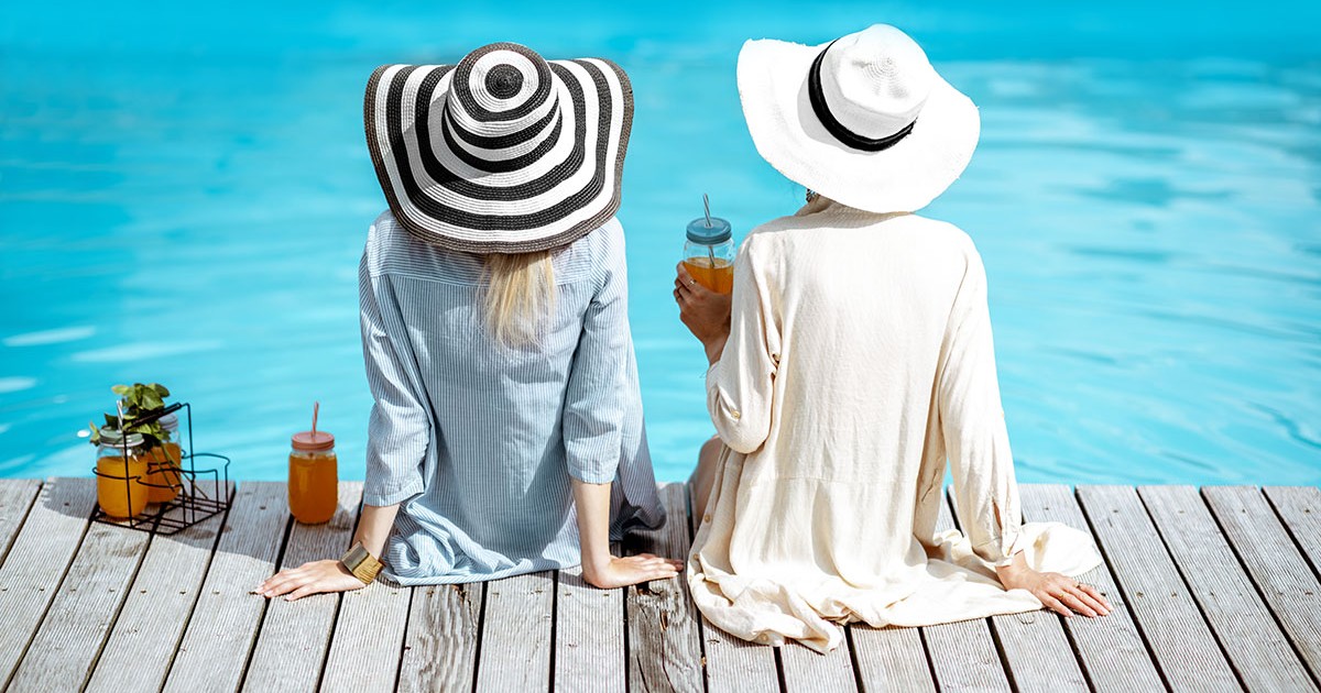 Două femei se relaxează la mare în fața unei ape albastre purtînd pălării.