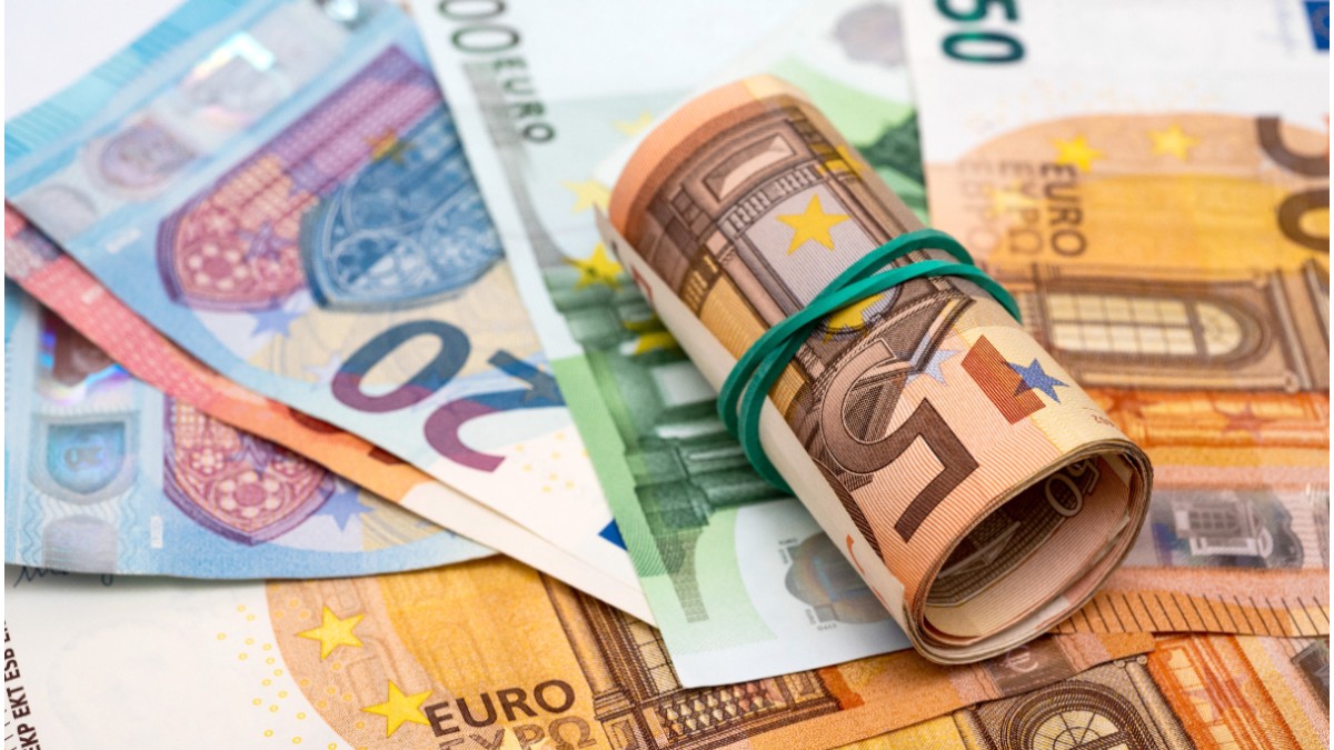 În imagine sunt prezentate mai multe bancnote euro împrăștiate, având în centru un pachet strâns de bancnote rulate și prinse cu un elastic de culoare verde