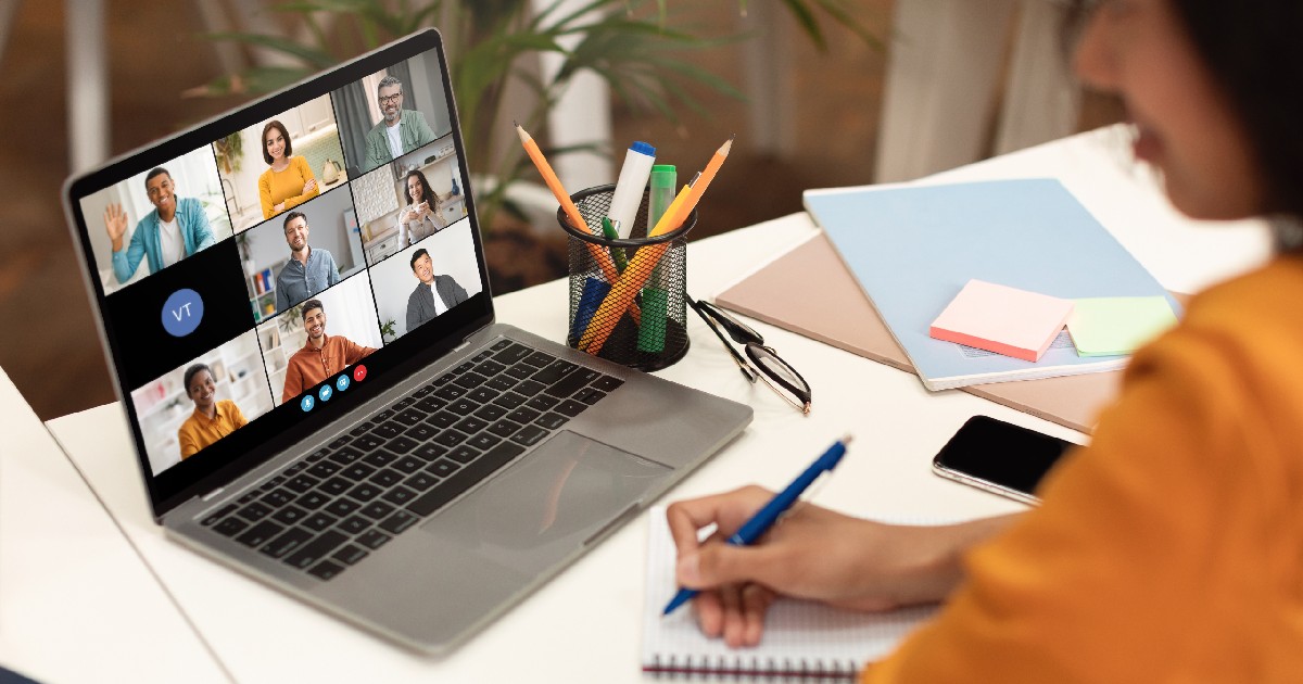 O femeie stă la un birou cu un laptop deschis, pe ecranul căruia se afișează o videoconferință cu mai mulți participanți. Ea ia notițe într-un caiet cu un stilou albastru, concentrându-se asupra ecranului.