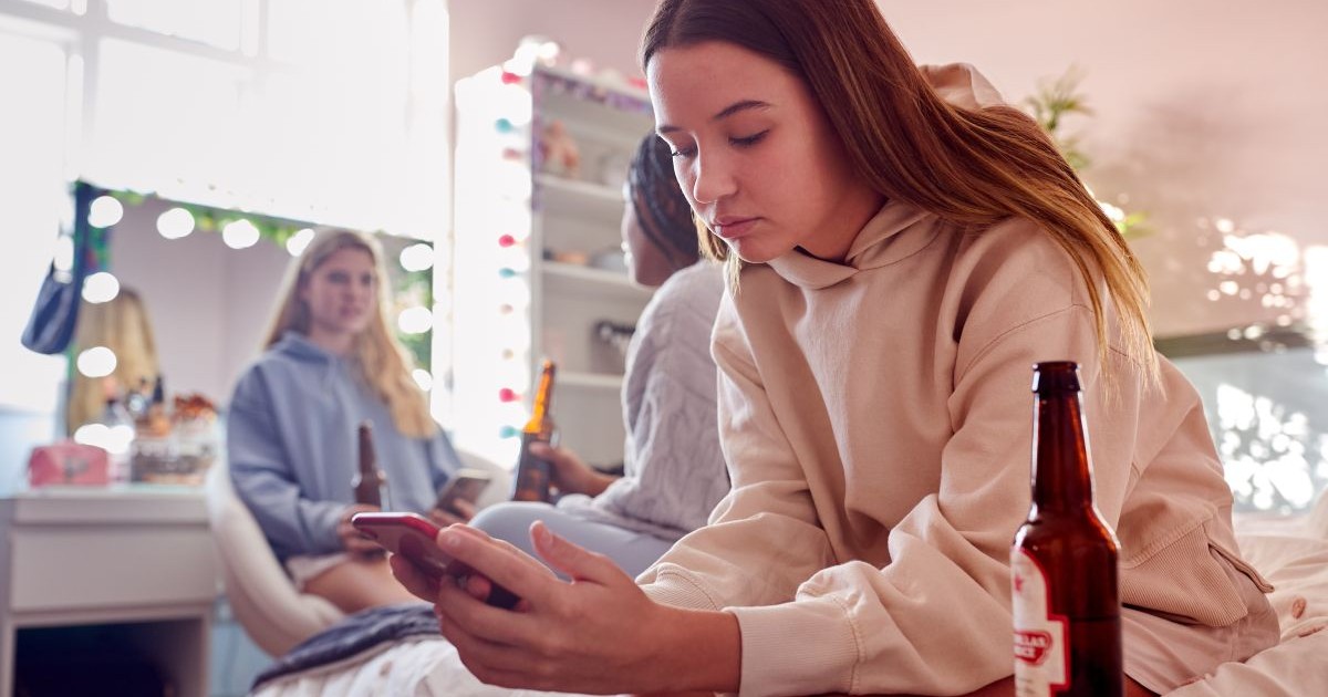 O adolescentă care stă pe pat, îmbrăcată cu un hanorac și pantaloni scurți, se uită în telefon și are lângă ea, pe noptieră, o sticlă de vere