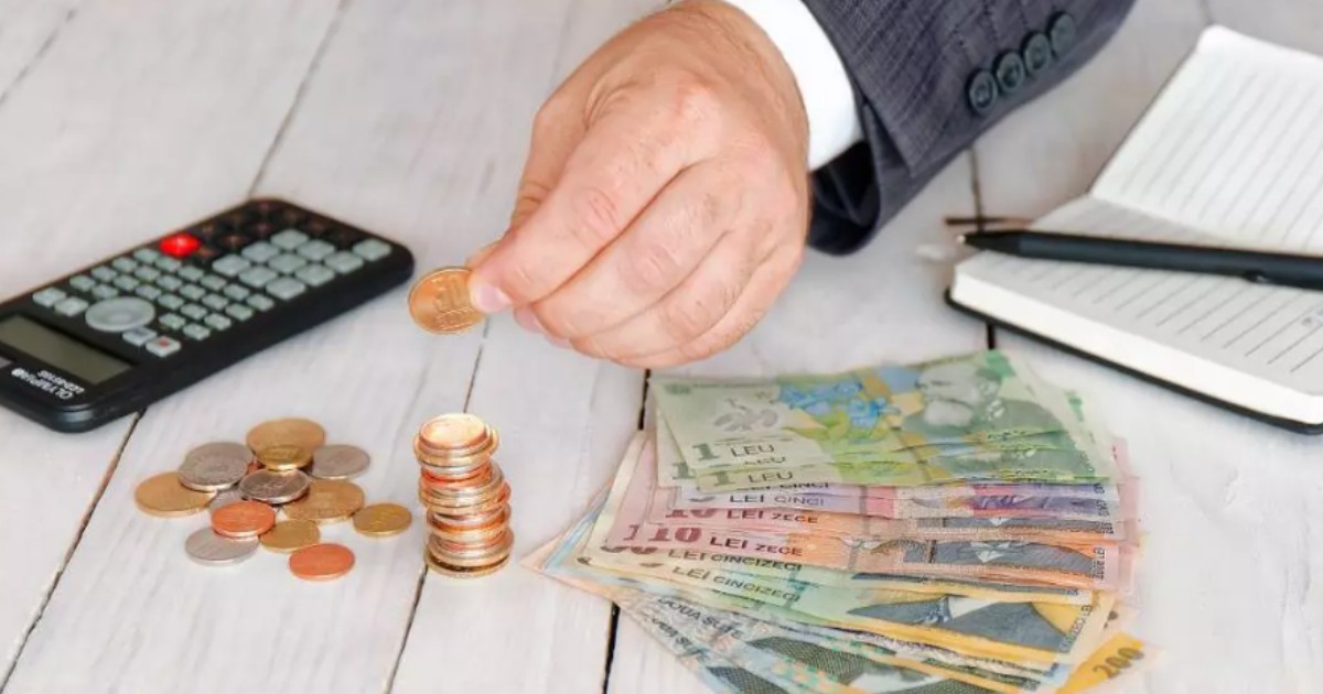 Mâna unui bărbat care pune o monedă pe o masă unde sunt mai mulți bani, o agendă, un pix și un calculator