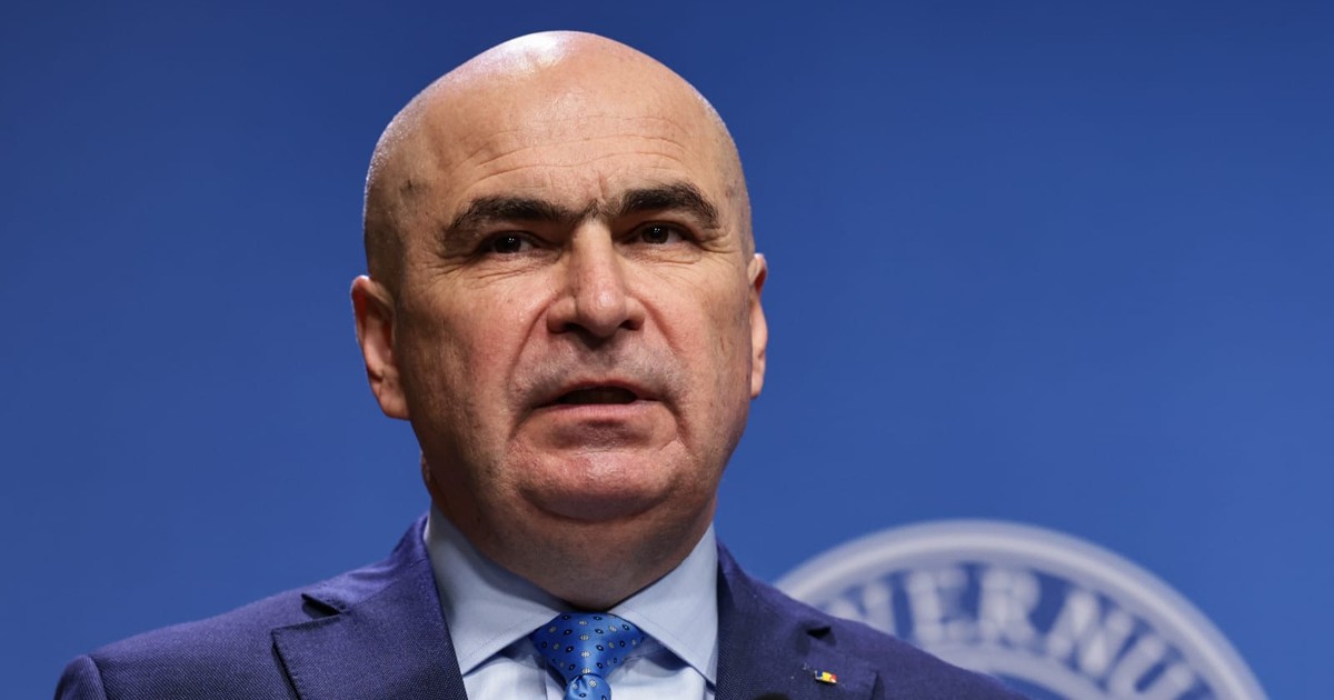 Premierul Ilie Bolojan, intr-un costum albastru, camasa belu deschis si caravata albastra, tine o conferinta la guvern despre sistemul de sanatate. in spate, pe un panou, se poate vedea o parte din logo-ul guvernului