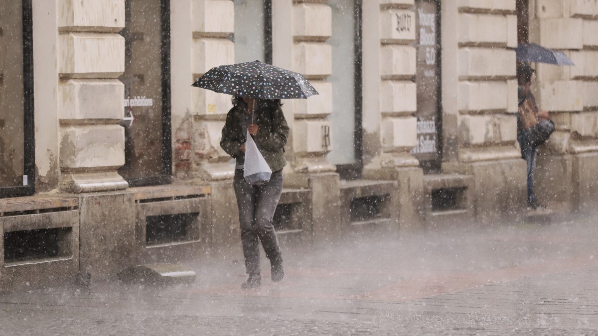 O persoană adăpostindu-se sub o umbrelă cu buline în timp ce merge pe trotuar pe o ploaie torențială, lângă fațada unei clădiri istorice.
