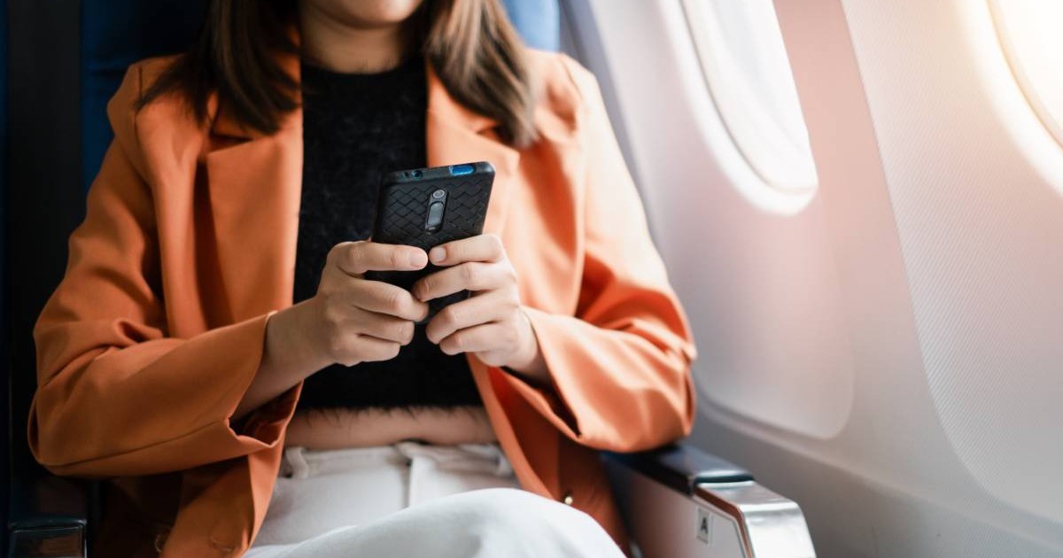 O femeie care stă cu telefonul în mână în avion și folosește internetul, poartă un palton portocaliu și pantaloni albi.