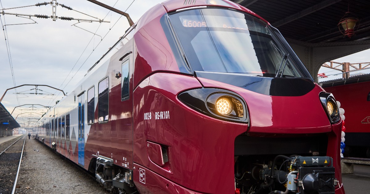 Imaginea prezintă un tren modern de călători, de culoare vișinie și albă, aflat într-o gară, pregătit pentru plecare sau sosire. În prim-plan se observă partea frontală aerodinamică a ramei electrice, iar în fundal sunt vizibile liniile de cale ferată, peroanele și infrastructura stației.