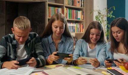 Patru elevi folosesc telefoane mobile, în timp ce sunt așezați la o masă acoperită de cărți și caiete deschise, in spatele lor se vede o bibliotecă