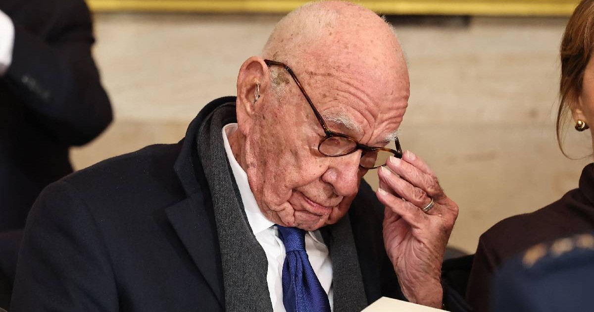 Magnatul Rupert Murdoch își ține ochelarii cu mâna în timp ce citește un document