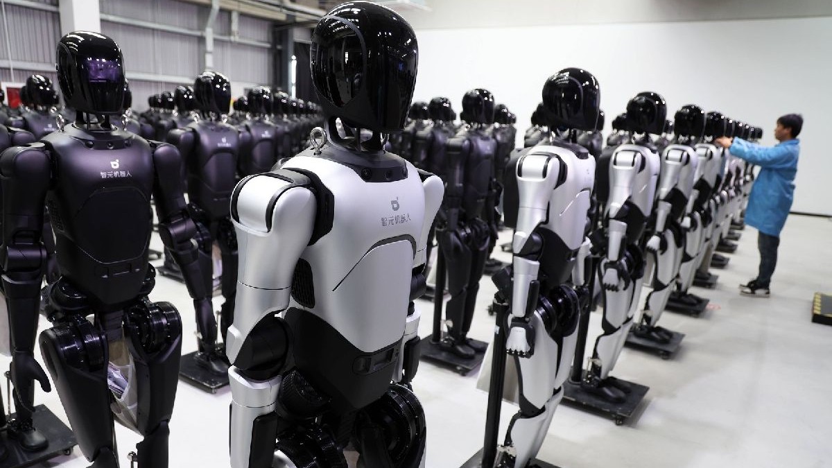 Rânduri de roboți umanoizi în alb și negru, așezați pe platforme. O persoană îmbrăcată într-o salopetă albastră reglează un robot.