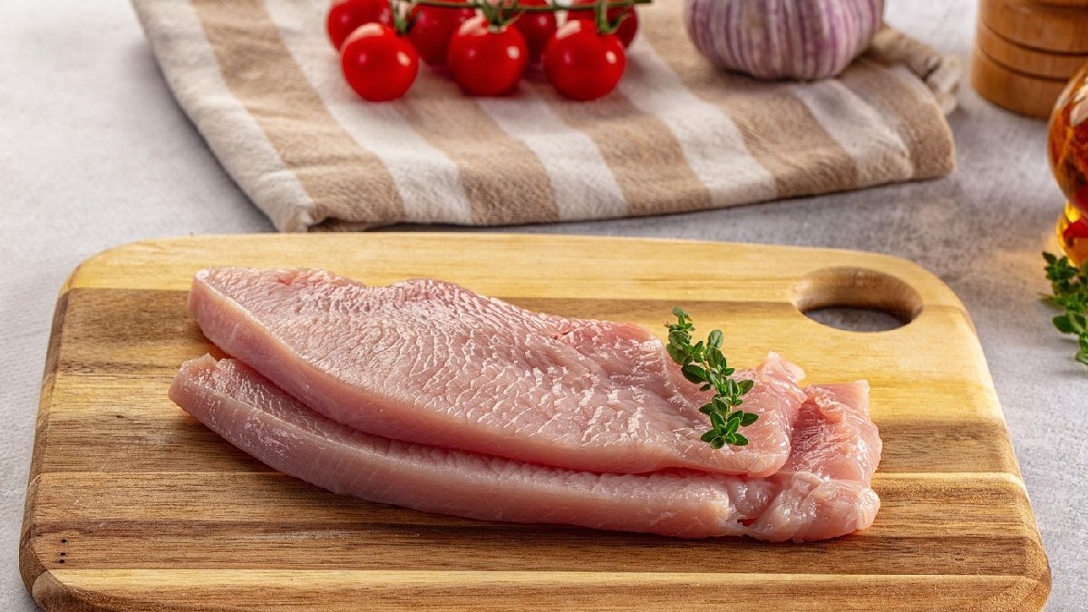 Filet de piept de curcan crud pentru gătit, pus pe un tocator de lemn, in spate se vad niste rosii și o căpățână de usturoi