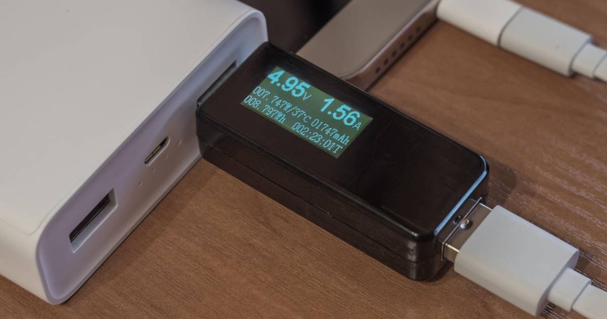 O baterie externă albă, conectată la un spartphone prin intermediul unui device de măsură a volților și amperilor.
