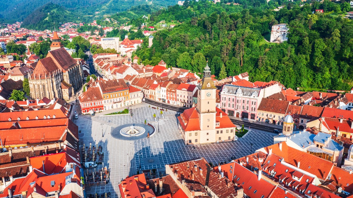 Imaginea surprinde o vedere aeriană a centrului istoric al Brașovului, cu Piața Sfatului, Casa Sfatului și Biserica Neagră în prim-plan. În fundal se întind dealurile împădurite care înconjoară orașul, oferind un contrast spectaculos cu acoperișurile roșiatice ale clădirilor istorice.