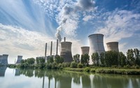 Termocentrala Turceni, cu un lac în față, turnurile uriașe pe fundal pe care iese fum și cerul albastru cu nori albi