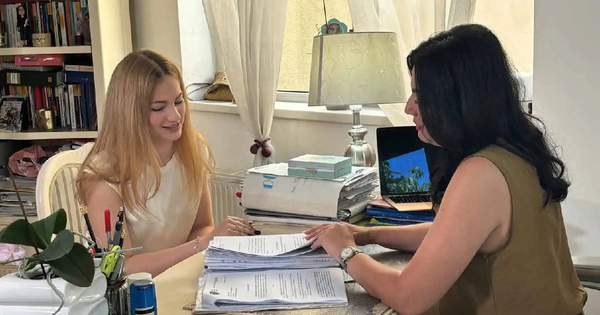 O tânără blondă, cu bluză alba, sta asezata in fata unei femei brunete, cu bluza kaki, la un birou, si se uită pe niște documente