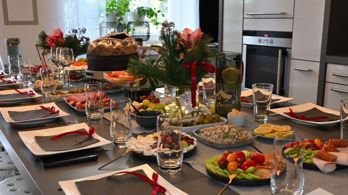 Masa de Crăciun este aranjată elegant, cu farfurii, pahare și șervețele decorative, într-un decor modern și luminos. Pe masă se află numeroase preparate festive, platouri cu aperitive, fructe și decorațiuni de sezon care creează o atmosferă caldă și primitoare.