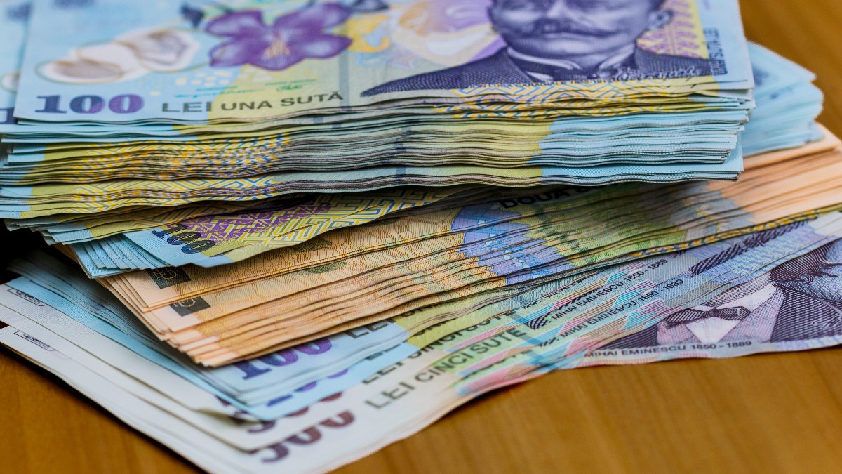 Imaginea prezintă mai multe teancuri de bancnote românești, de diferite valori, așezate pe o suprafață de lemn. Culorile distincte și portretele de pe bancnote sugerează sume importante de bani și un context financiar sau economic.