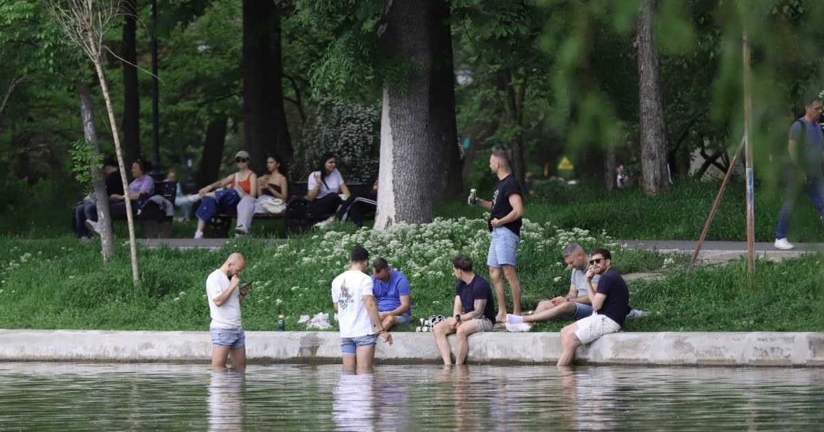 Doi băieți stau în picioare în apă, alți trei stau pe bordura lacului și cu picioarele în apă, un altul stă în picioare pe iarbă cu o doză în mână, iar în spate se mai văd câteva tinere și alți oameni care sunt pe bancă, în parcul Cișmigiu, București, 25 aprilie 2025