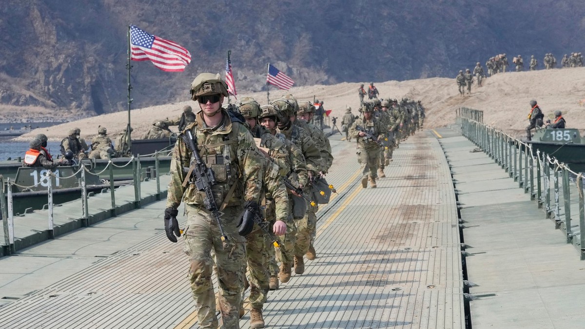 O coloană de soldați americani în echipament complet de luptă, cu steaguri ale SUA arborate, traversează în șir indian un pod ponton peste un râu lat, flancat de bărci militare și maluri aride.