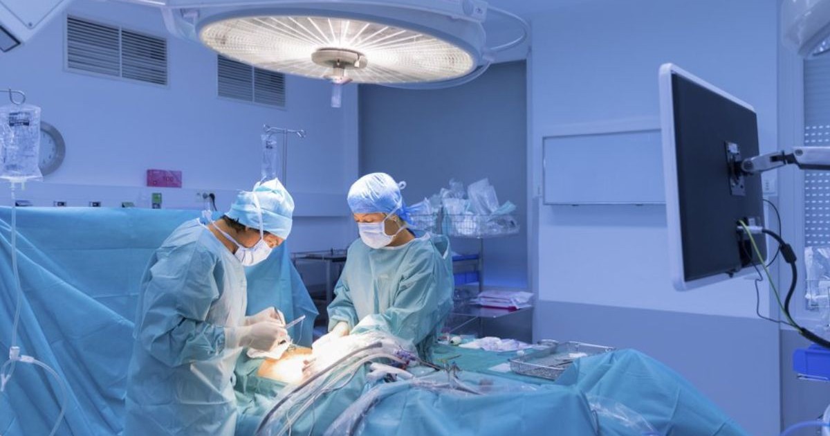 doi chirurgi in sala de operație, aplecati asupra unui pacinet, intr-o lumina albastra, cu o lapa mare deasupra mesei de operație.