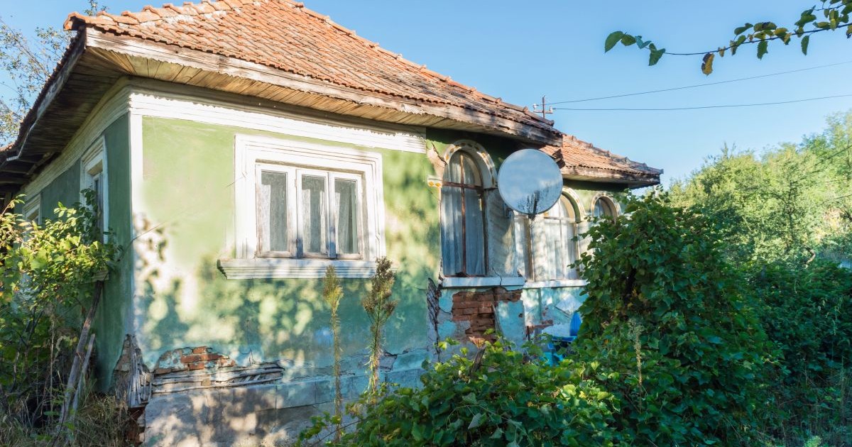O casă veche de la sat, vopsită verde deschis, cu tâmplărie veche albă, cu câteva bucăți de zid căzute, are în jur vegetație