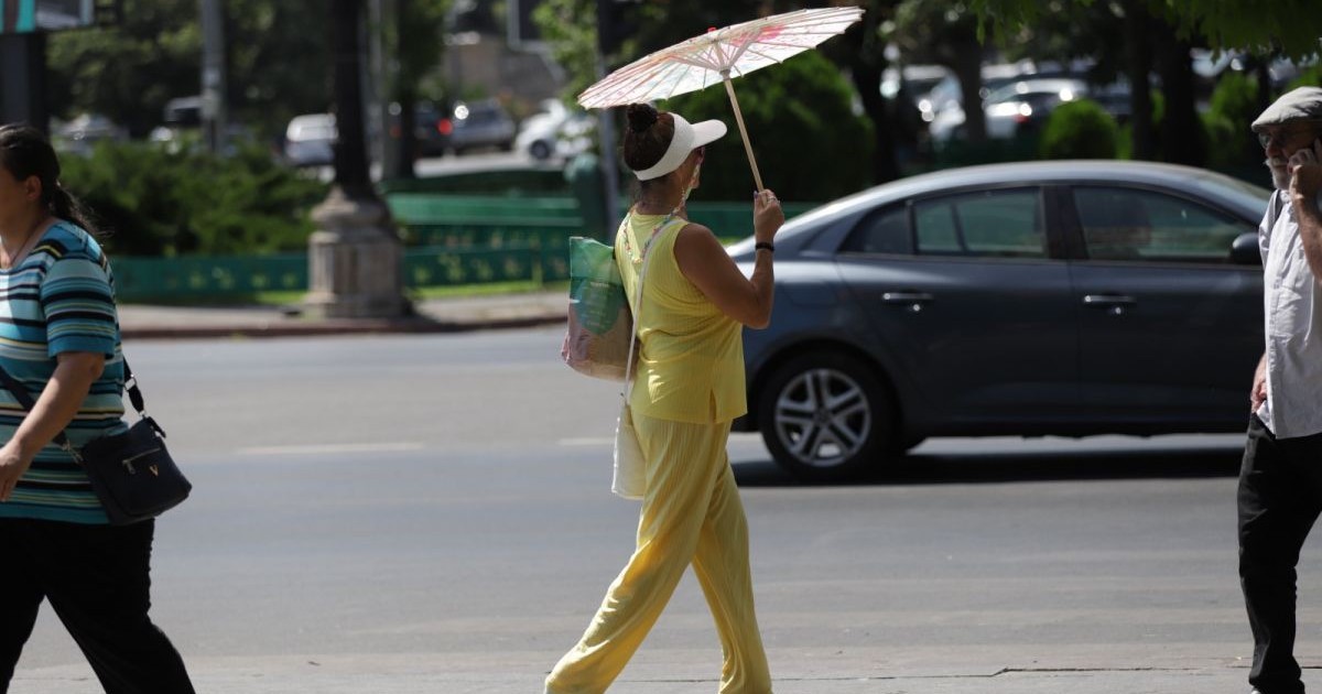În centrul imaginii este o femeie îmbrăcată în galben, cu o șapcă albă și umbrelă de soare deasupra capului, în dreapta este un bărbat care vorbește la telefon, iar în stânga o femeie. În spate, se vede o mașină pe stradă, București, 28 iunie 2025
