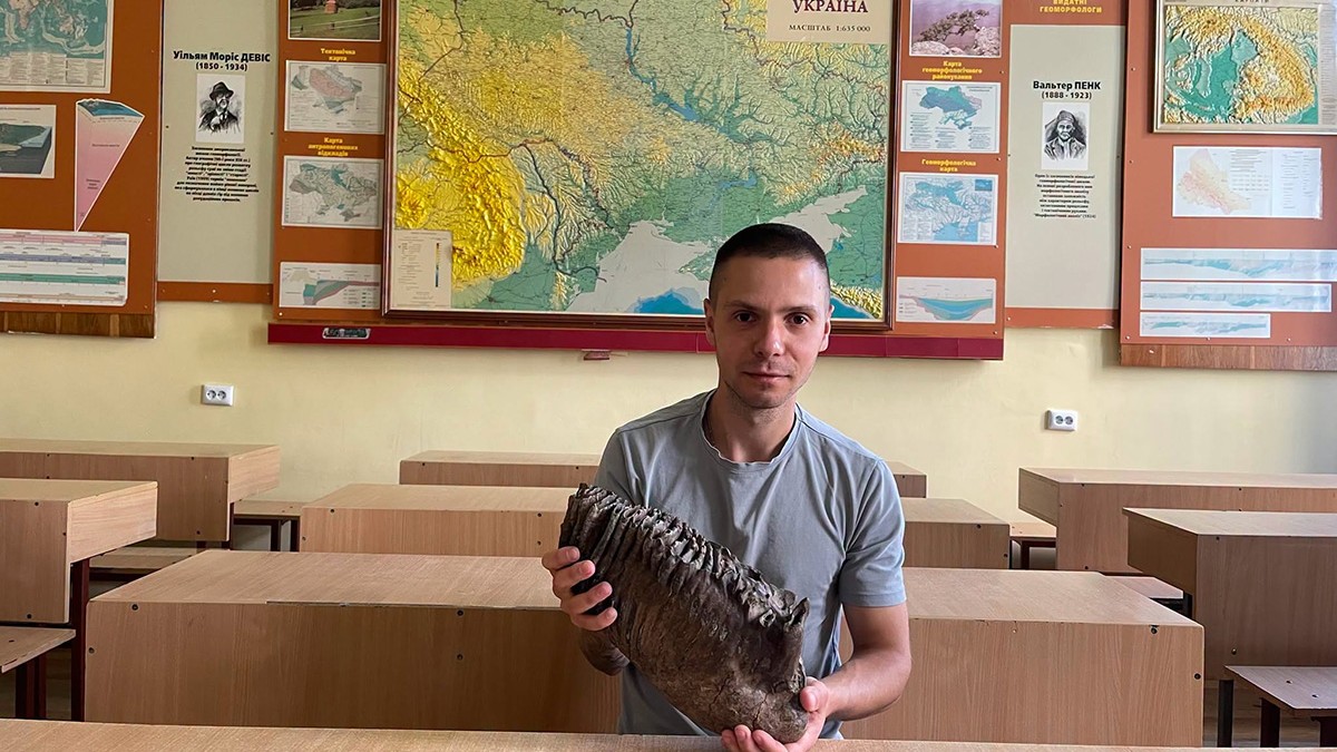 Volodimir Proț cu dintele de mamut într-o clasă iar în spatele său se află o hartă.