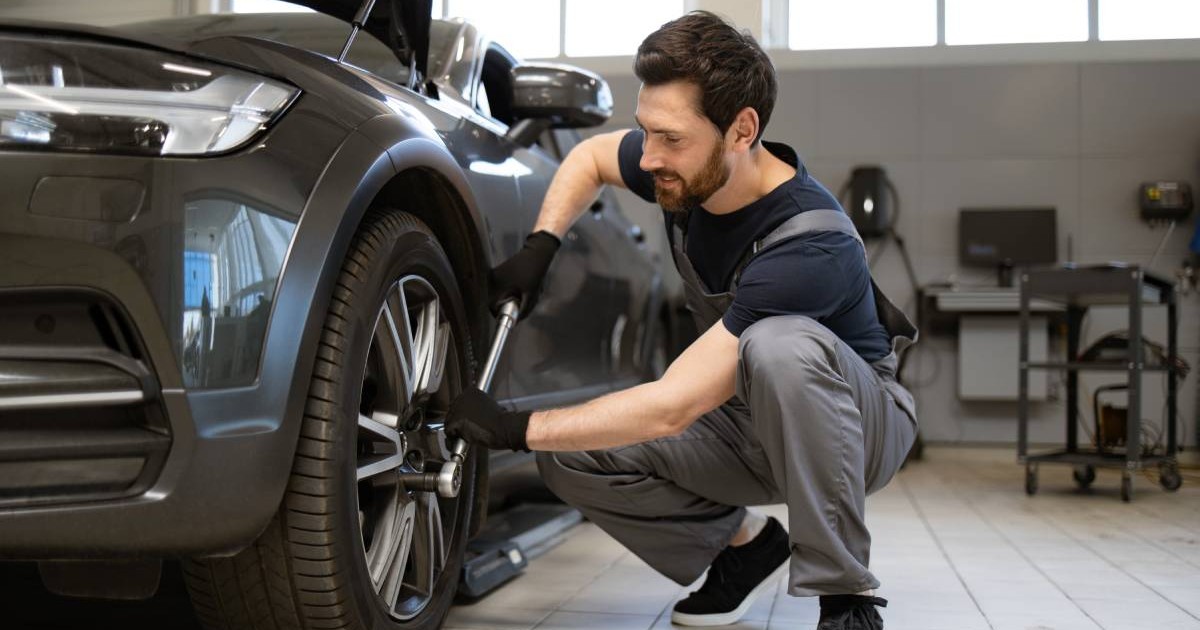 Un mecanic auto îmbrăcat cu salopetă gri și tricou negru, în timp ce strânge prezoanele roții unei mașini negre