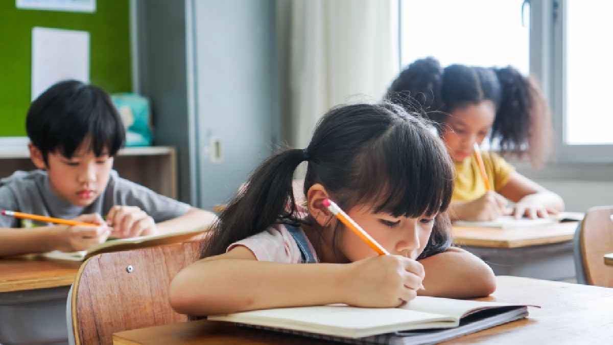 Elevă stând în bancă și scriind în caiet cu creionul, studiind, educație, învățare. Copii asiatici în clasă. Diversitate în rândul elevilor.