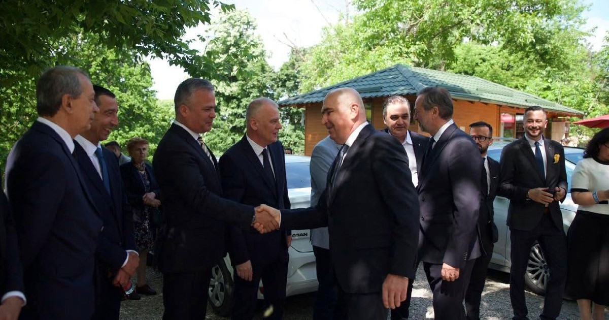 Adrian Veștea dă mâna cu Ilie Bolojan, într-o curte plinî de copaci, amândoi poartă costume închise la culoare