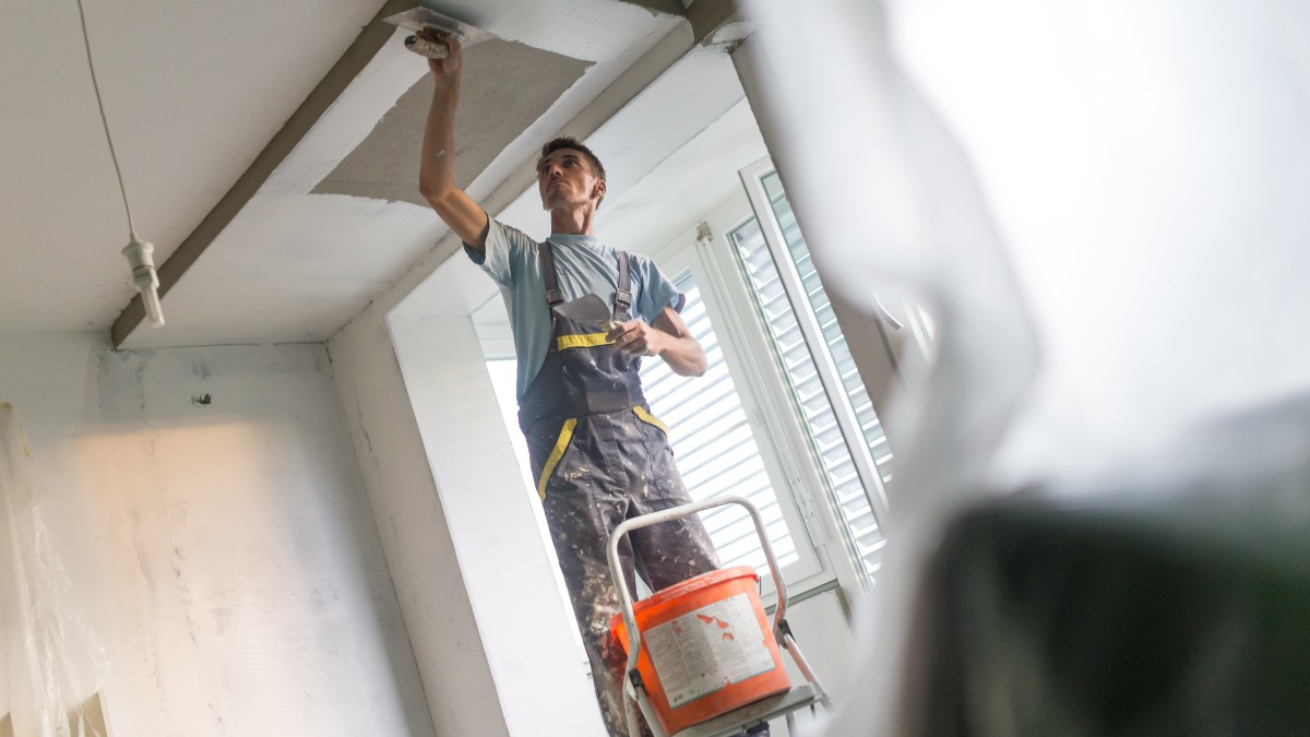 Imaginea prezintă un muncitor zugrav îmbrăcat în salopetă care aplică glet pe tavanul unei camere aflate în renovare, folosind o gletieră.