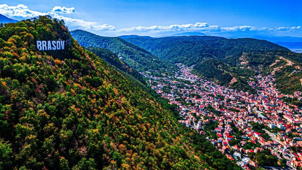 Imagine aeriană cu orașul Brașov în care se vede muntele pe care este montat semnul cu numele orașului Brașov și case aflate la poalele muntelui