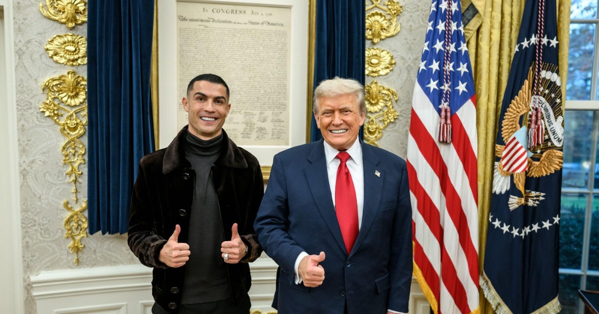 Fotbalistul Cristiano Ronaldo și omul de afaceri Donald Trump stau împreună într-un cadru oficial, cel mai probabil la Casa Albă, ambii zâmbind și arătând gestul de „thumbs-up” cu mâna. În spatele lor se observă drapelul american și un document înrămat, alături de perdele din catifea albastră și decorațiuni aurii.
