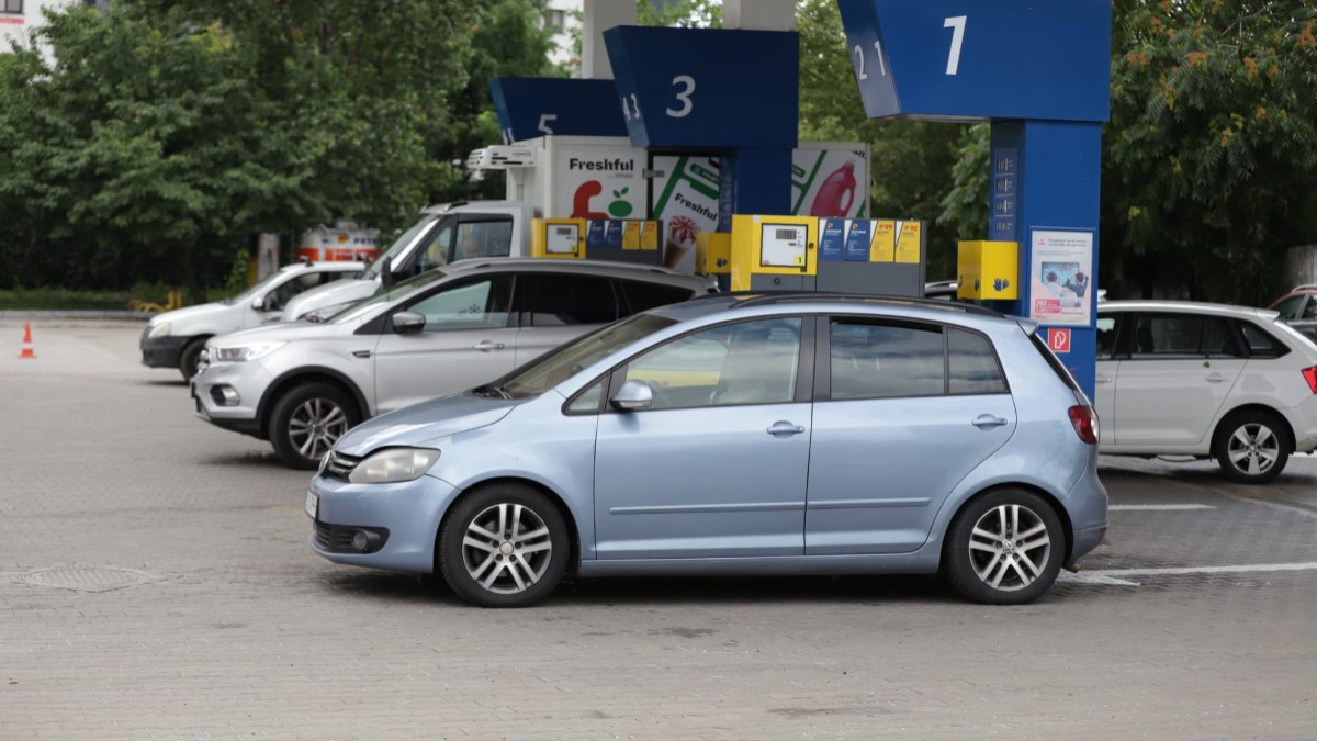 Imaginea prezintă mai multe mașini, printre care un Volkswagen de culoare albastru deschis, staționate la pompele unei benzinării.