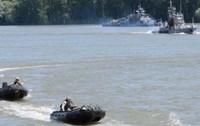 Un exercițiu militar pe apă, în care mai multe bărci pneumatice rapide ghidate de militari înaintează pe un fluviu, având nave militare de patrulare în fundal.