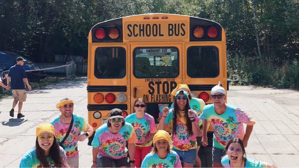 9 americani imbracati in tricouri colorate pozeaza langa un autobuz galben scolar din SUA