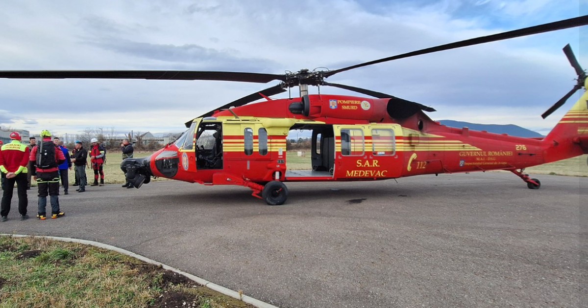 Un elicopter de salvare de tip Black Hawk, vopsit în culorile roșu și galben, cu însemnele GUVERNUL ROMÂNIEI și S.A.R. MEDEVAC, staționat pe o pistă, având alături membri ai echipajului în uniforme de intervenție.