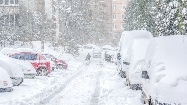 O stradă dintre blocuri acoperită de un strat gros de zăpadă în timpul unei ninsori abundente, cu numeroase autoturisme parcate pe margine și o persoană care merge pe aleea îngustă curățată.
