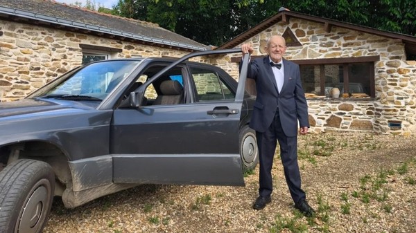 Un bărbat în vârstă, îmbrăcat elegant în costum cu papion, stând lângă portiera deschisă a unei mașini, în fața unei case din piatră.