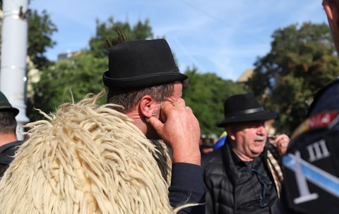 Un bărbat îmbrăcat în cojoc tradițional de blană și cu pălărie neagră, dus cu mâna la ureche, aflându-se printre alți protestatari și un jandarm în timpul unei manifestații stradale.