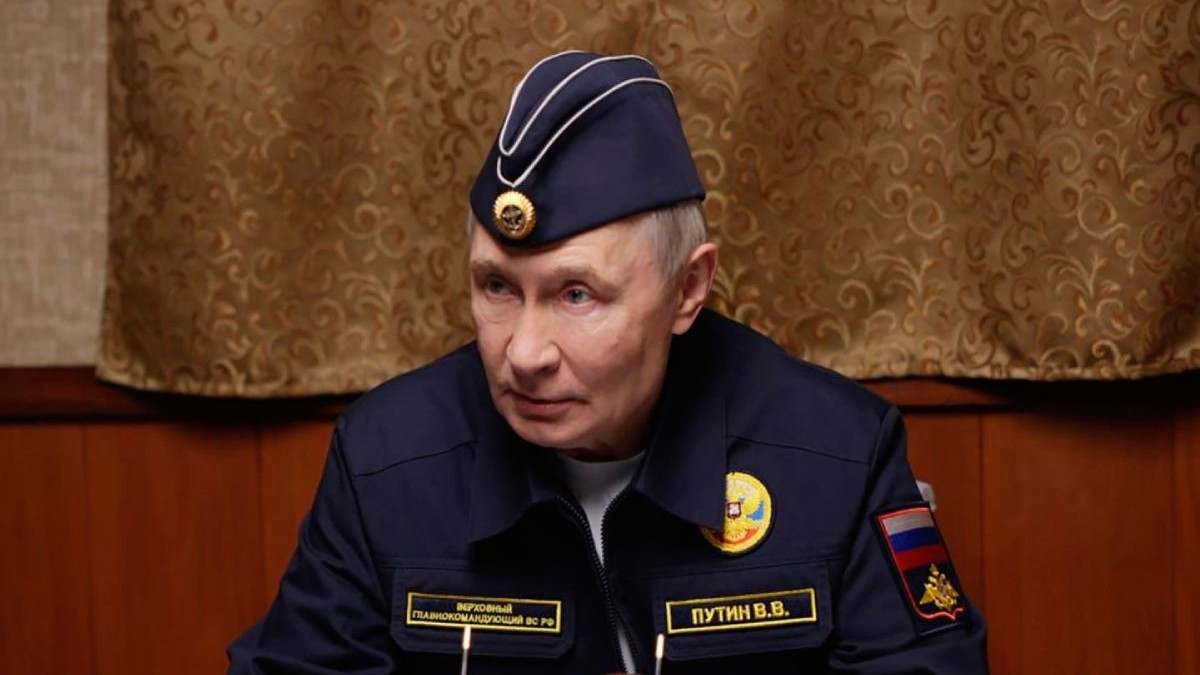 Vladimir Putin îmbrăcat într-o uniformă militară de Marină, în timp ce stă pe scaun și are pe fundal o perdea de culoare aurie